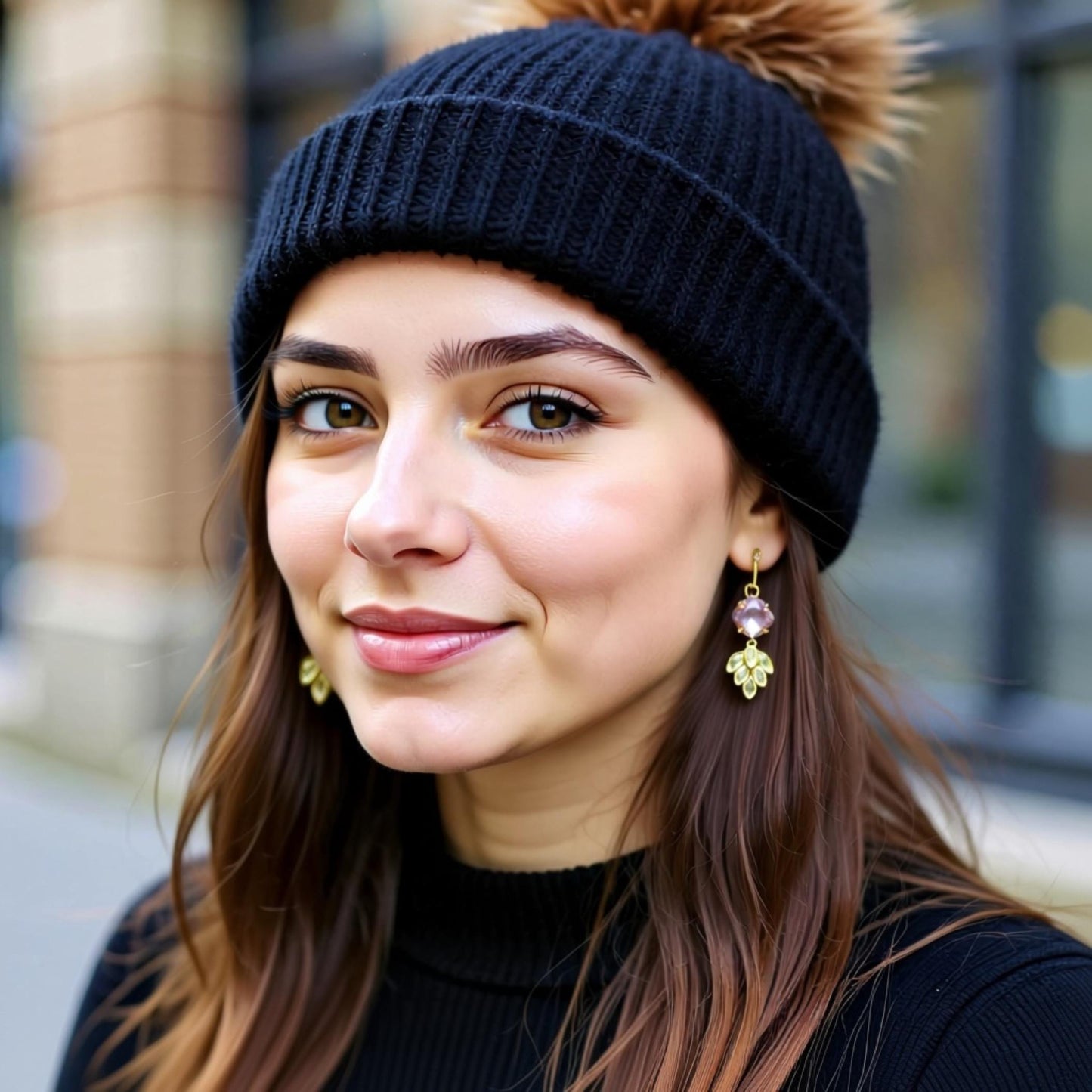 A young woman with long brown hair wearing a black beanie and gold earrings, smiling at the camera.
