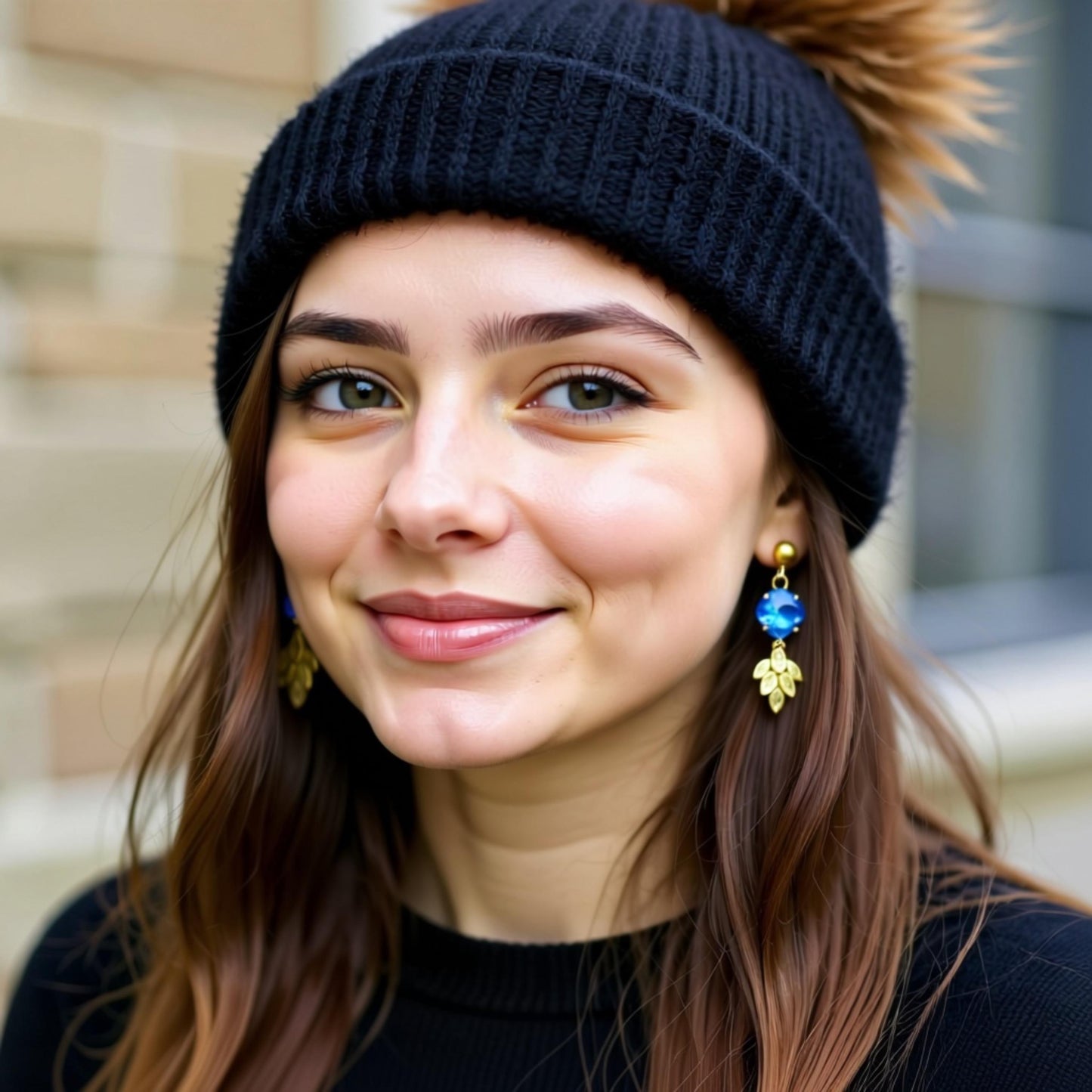 A young woman with long brown hair wearing a black beanie and gold leaf earrings, smiling at the camera.