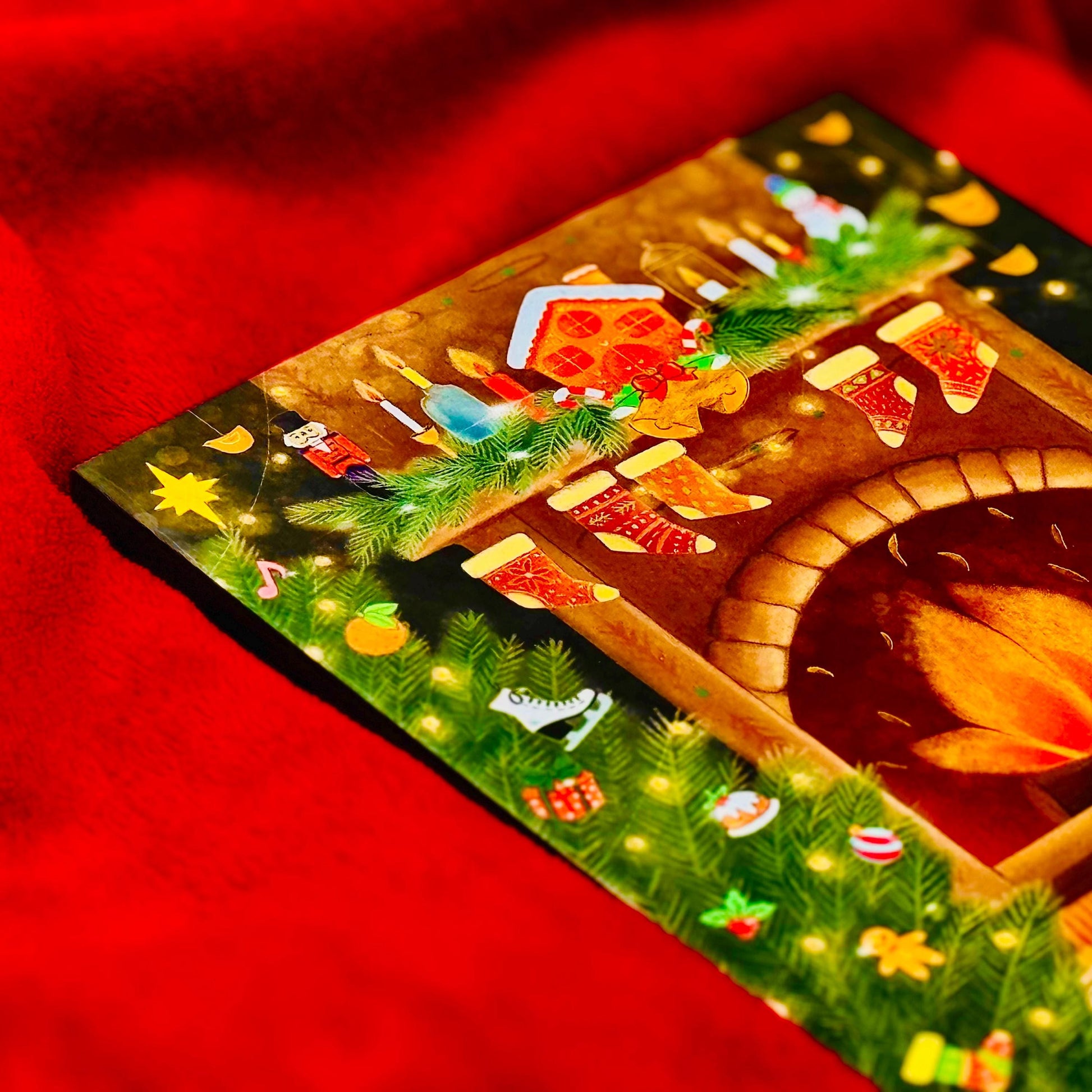 A colorful Christmas card with a fireplace, stockings, and other festive decorations is displayed on a red surface.