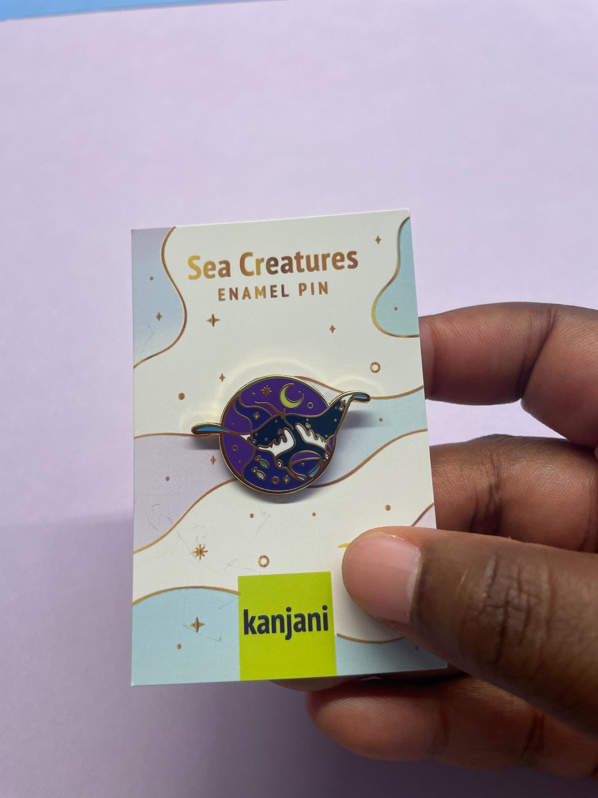 A hand is holding a pin with a purple and gold design, which appears to be a Sea Creatures pin.