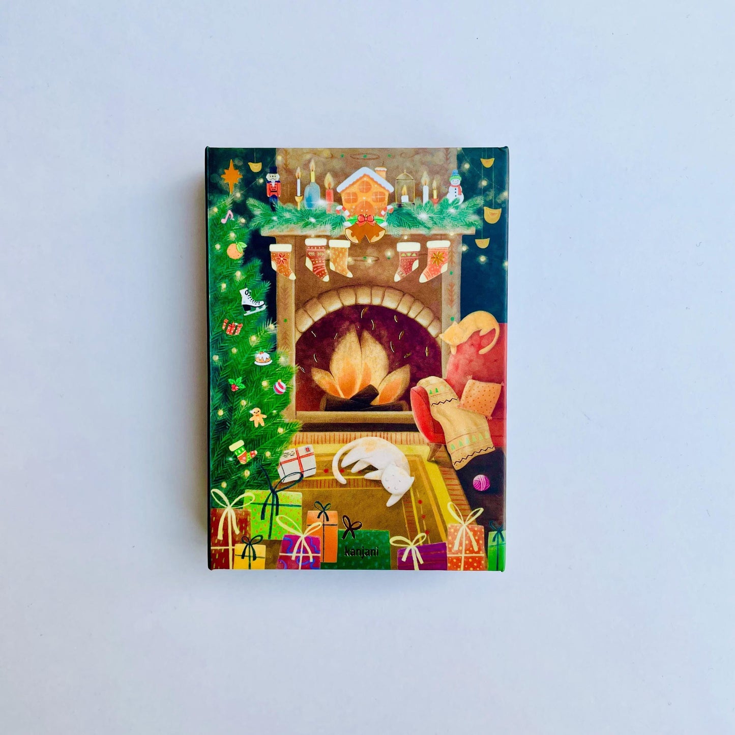 A colorful Christmas card depicting a cozy fireplace with a decorated tree, presents, and a cat curled up by the fire.