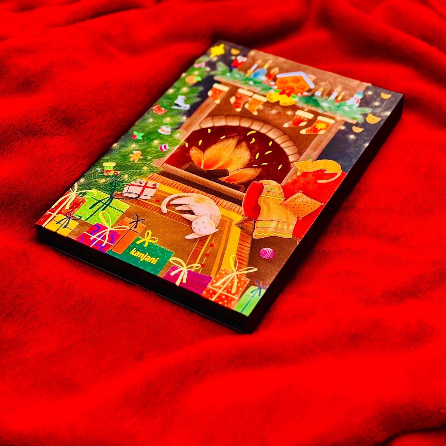 A colorful Christmas card with a fireplace, presents, and a cat is displayed on a red surface.