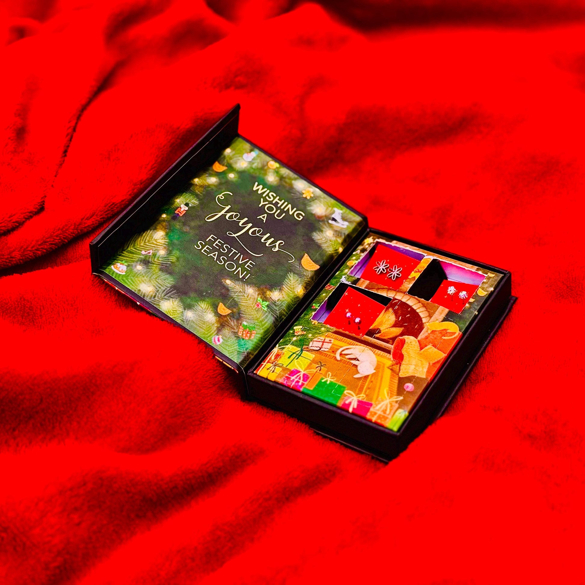 An open gift box with a festive design, placed on a red fabric surface.