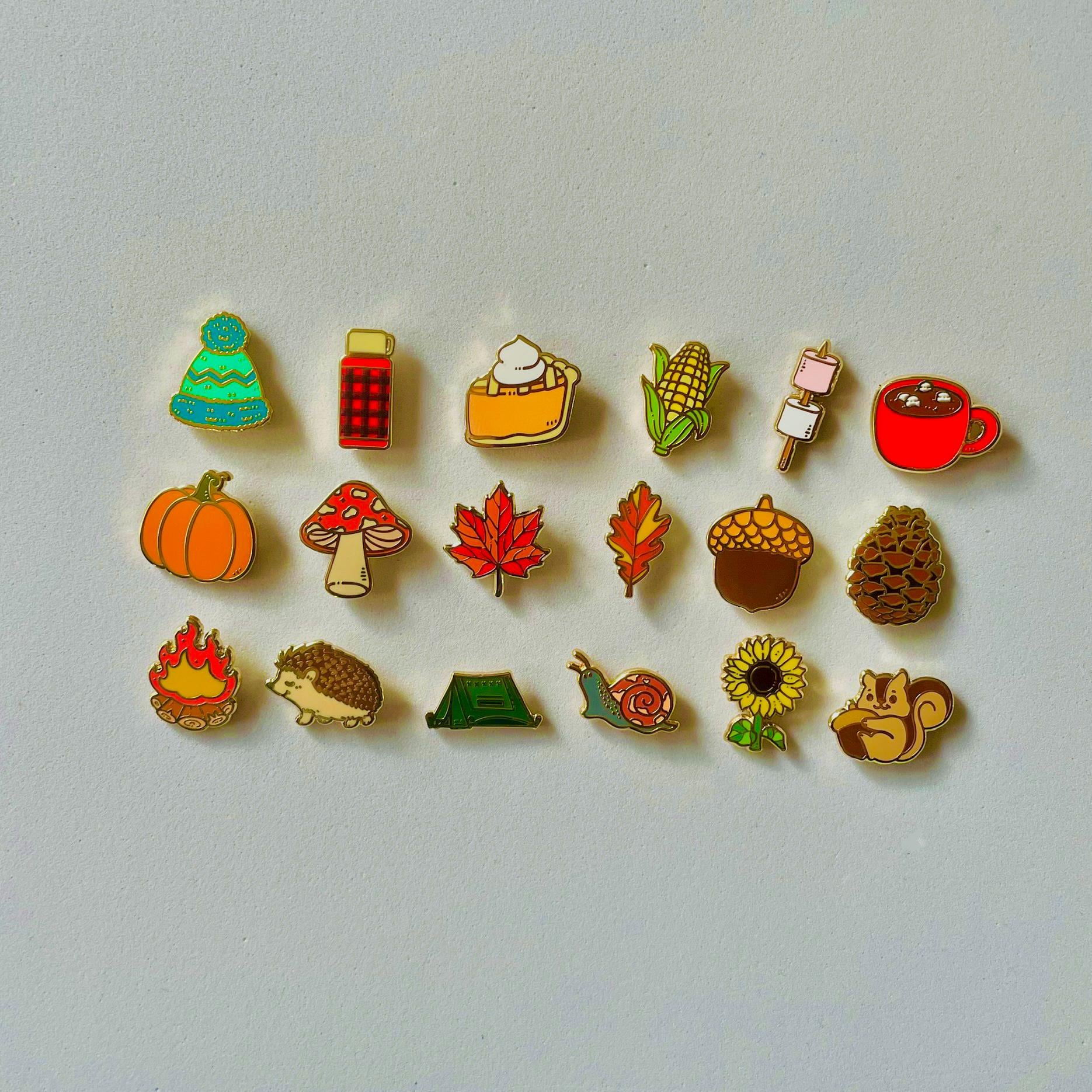 a collection of 16 colorful pin badges, each depicting a different autumn-themed object or symbol, such as a pumpkin, maple leaf, pine cone, mug, and more.