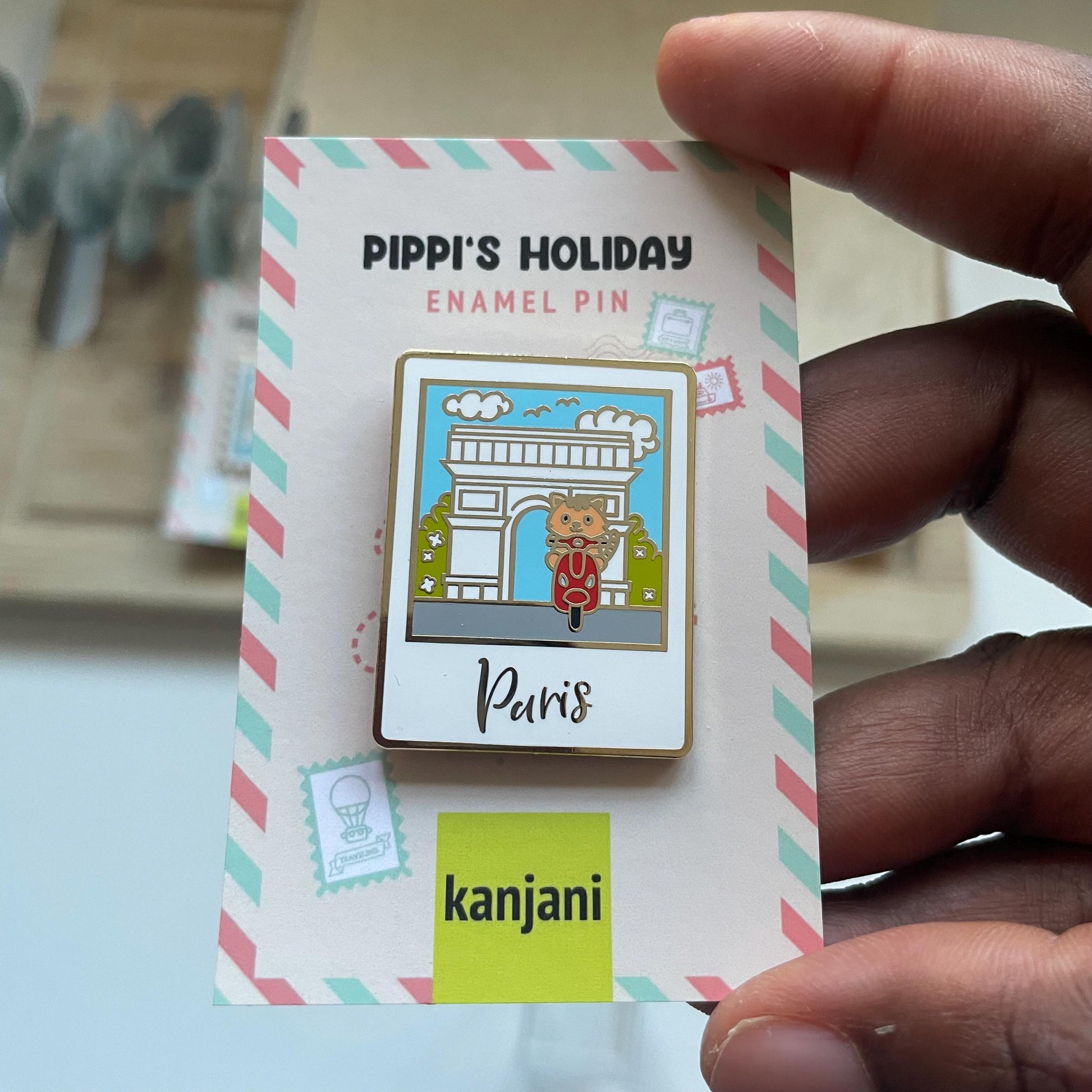 A person is holding a Pippi's Holiday Enamel Pin, which features a cartoon image of a dog riding a motorcycle. The pin is displayed on a card with a striped border and the word "Punis" written on it.