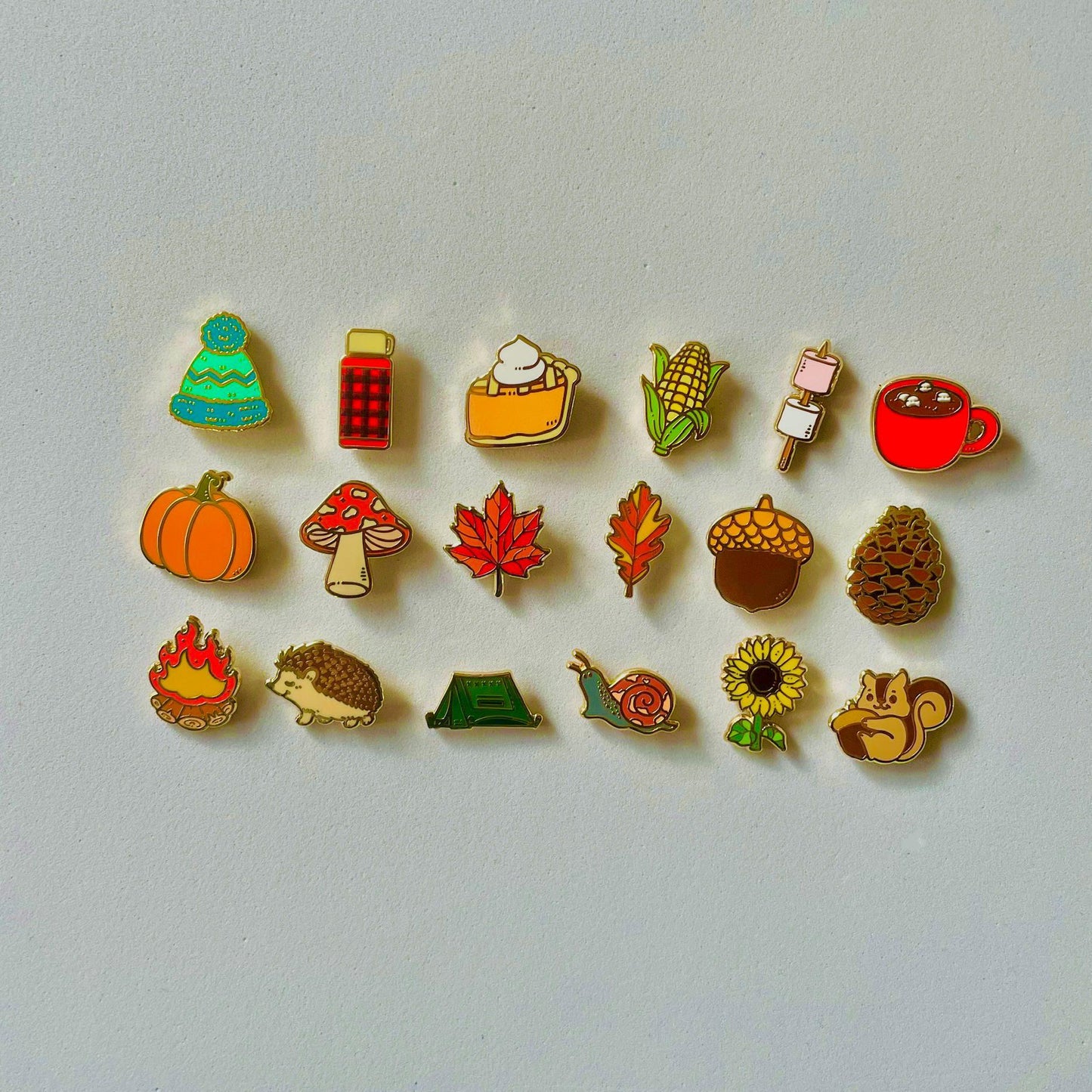 a collection of 16 colorful pin badges, each depicting a different autumn-themed object or creature, such as a pumpkin, hedgehog, squirrel, and more.