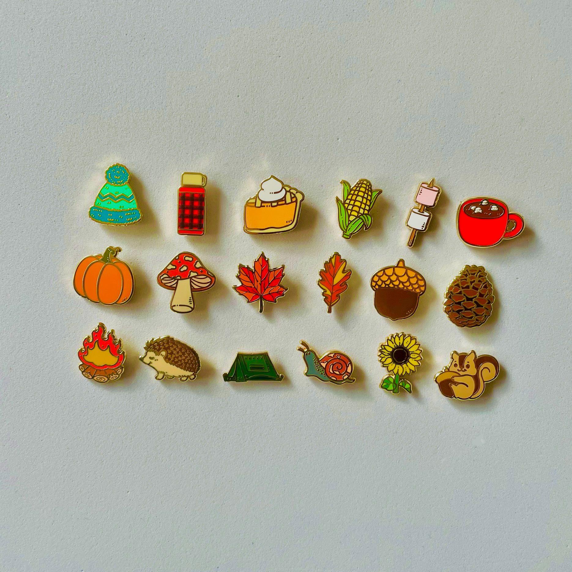 a collection of 16 colorful pin badges, each depicting a different autumn-themed object or creature, such as a pumpkin, hedgehog, squirrel, and more.