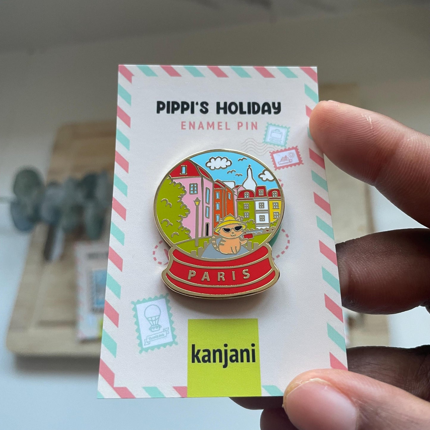 A person is holding a colorful enamel pin featuring a cartoon image of Paris, with a cat and a building in the background.