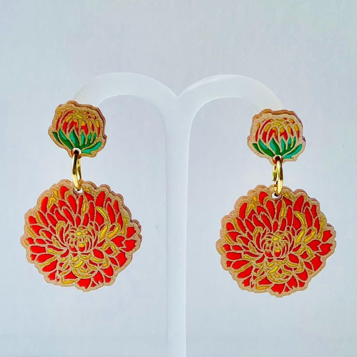 A pair of earrings with red and gold floral designs, hanging from a white stand.