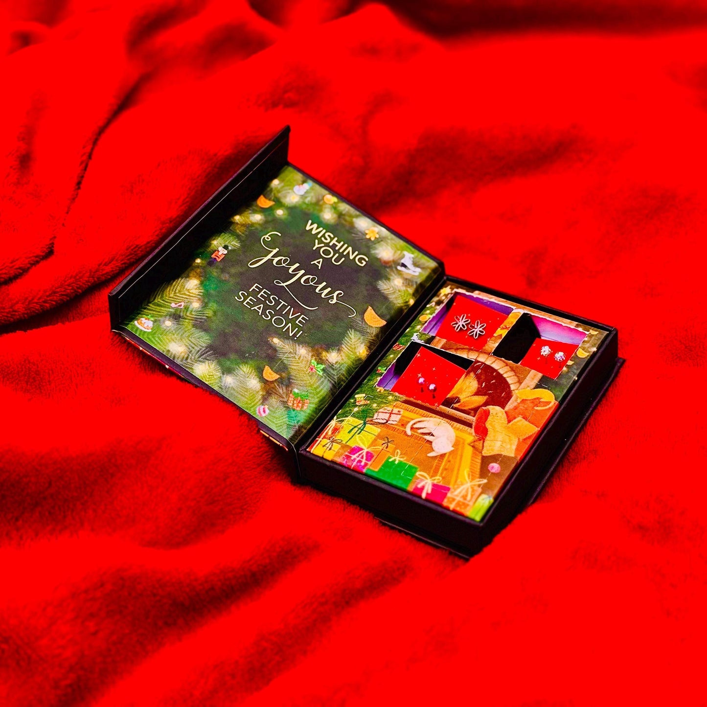 An open gift box with a festive design, placed on a red fabric surface.