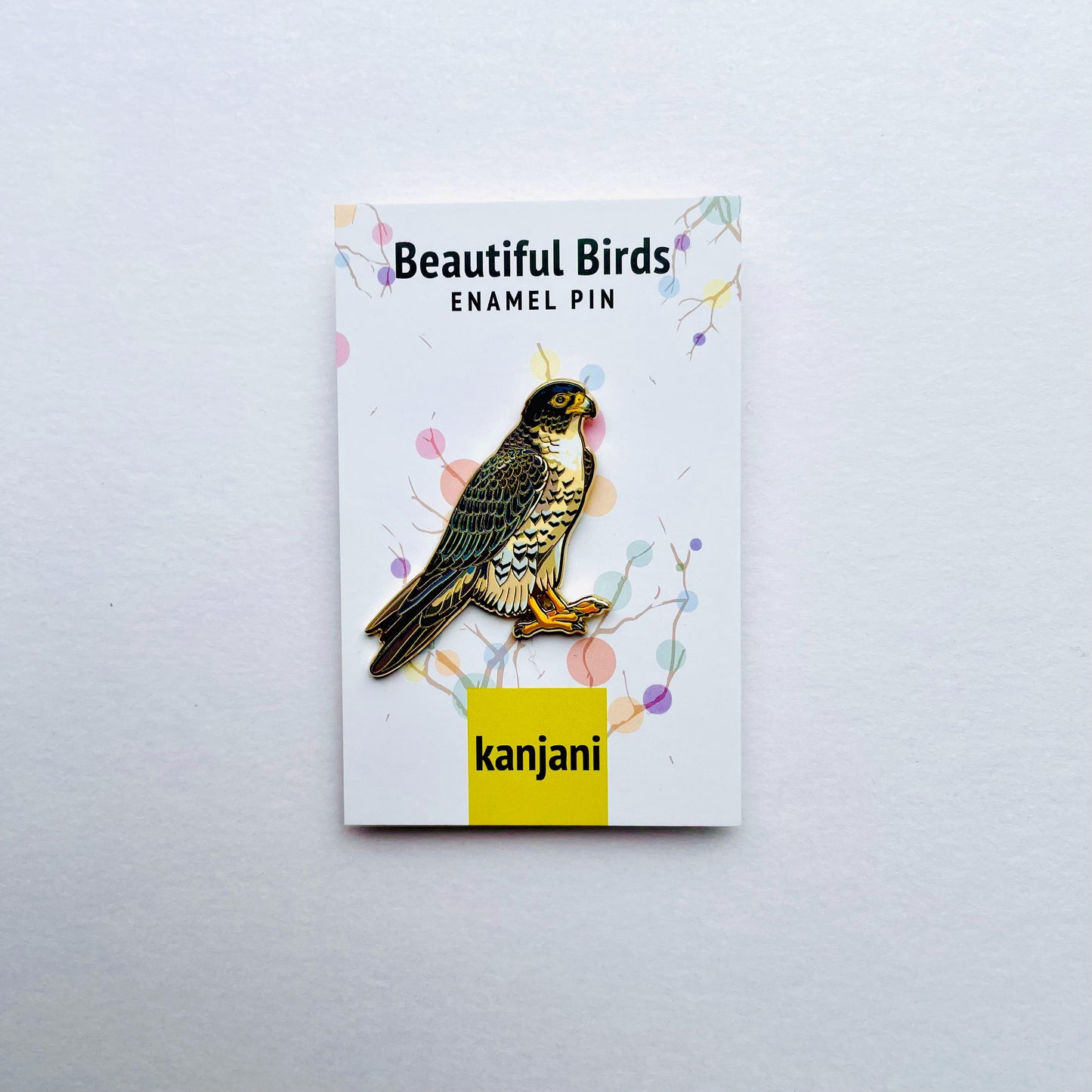 a beautiful bird enamel pin with a colorful background and a yellow label that reads 'kanji'.