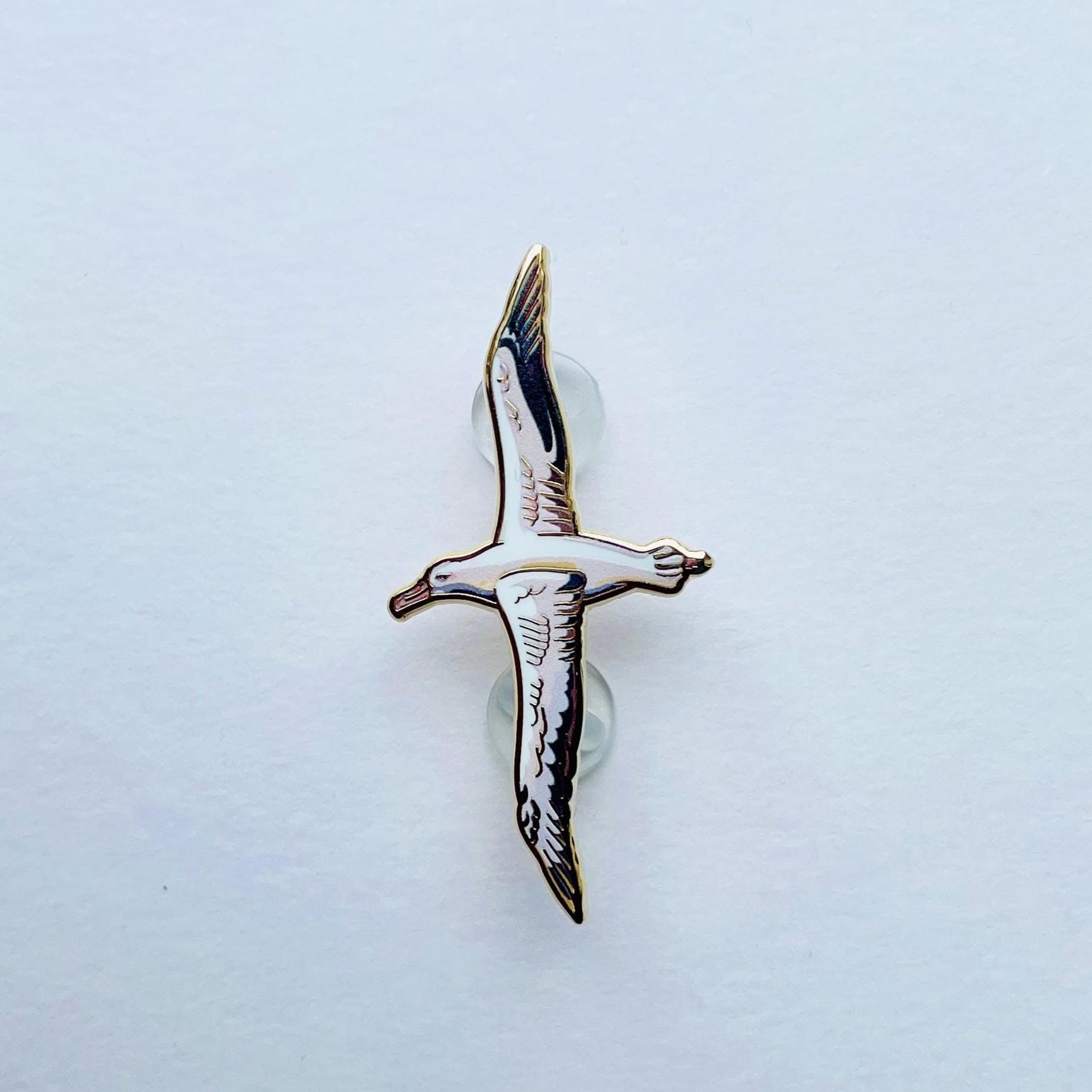 A single, detailed enamel pin depicting a bird in flight against a plain white background.